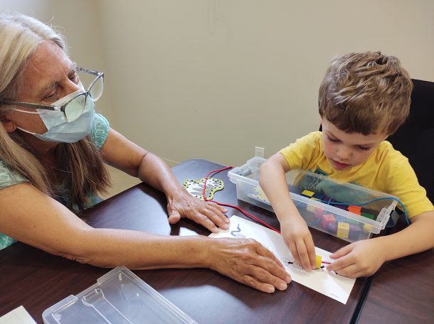 A woman holds a piece of paper on a table while a young boy places beads on top of it.