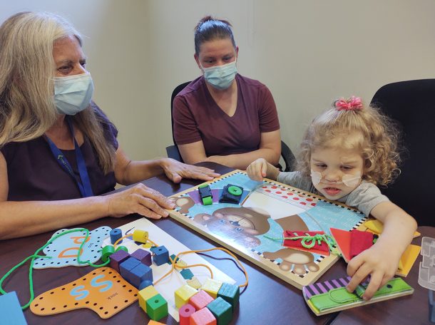 2 women watch as a young girl picks up a puzzle piece.