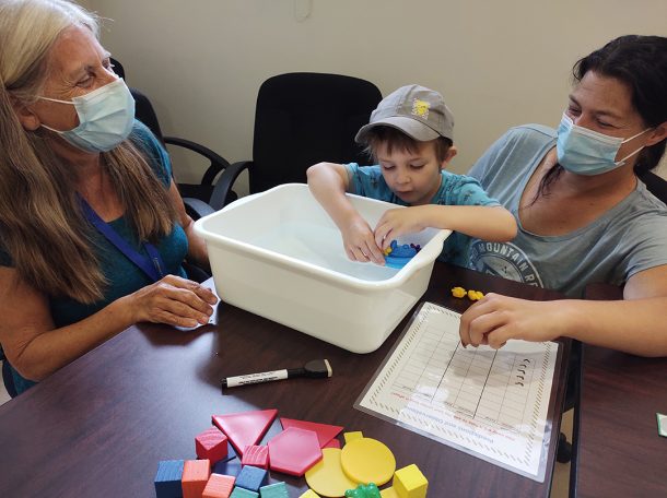 2 women watch as a young boy places a small plastic bear on a plastic boat floating in a small tub of water.