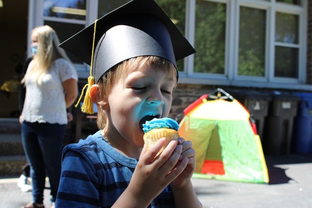 A young boy in a paper graduate cap takes a bite out of a cupcake covered with blue icing.