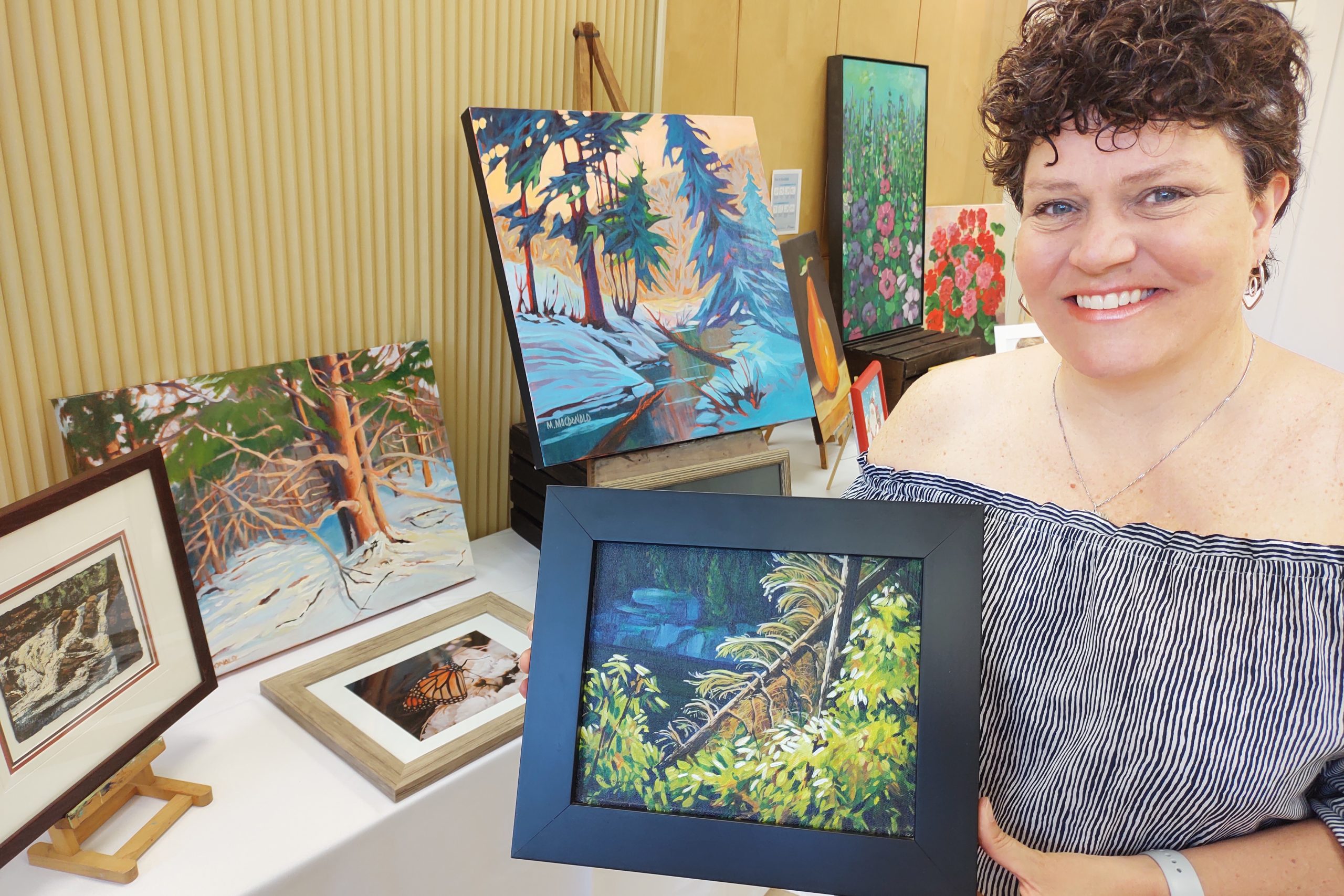 Smiling woman holds a small, framed landscape painting, while standing in front of a table filled with art.