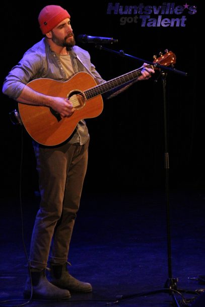 A man in an orange hat stands on stage singing into a microphone while playing a guitar.