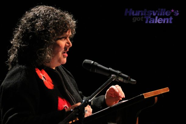 a woman stands at a podium on a theatre stage and talks into a microphone