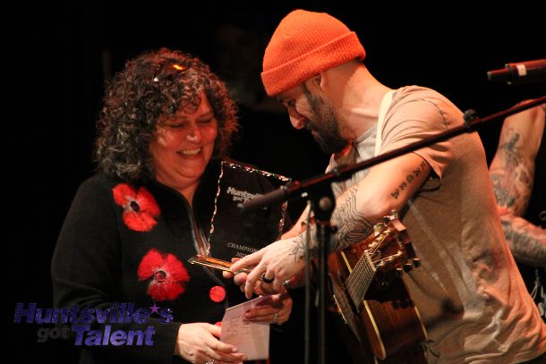 A smiling woman hands a smiling man in an orange hat a champion's award.