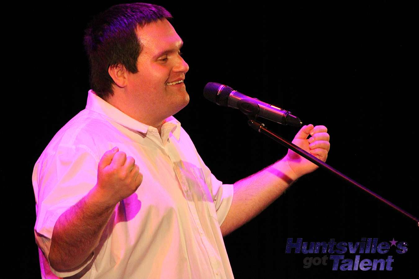 a young man in a white, collared shirt raises his hands with outstretched arms as he smiles and sings into a microphone
