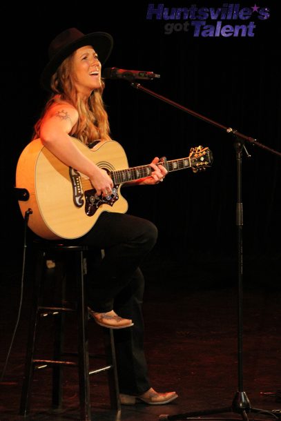 a young woman with blonde hair and a wide-brimmed hat seated on a tall stool sings into a microphone while playing a guitar.