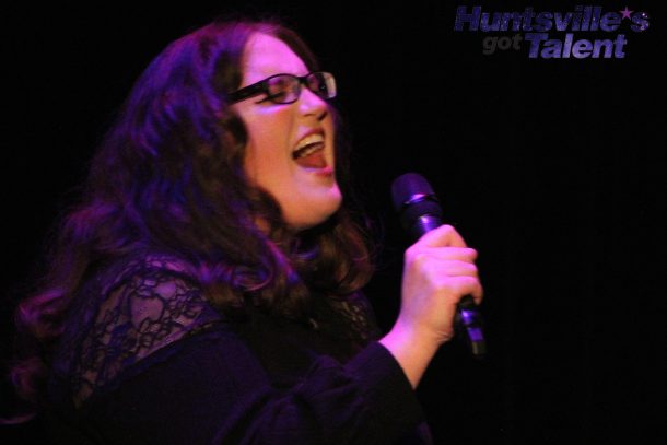 a woman with dark hair and glasses sings passionately into a microphone.
