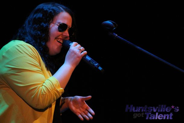 a woman in a yellow jacket and sunglasses sings into a microphone.