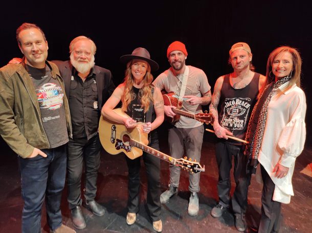 6 smiling people pose together for a photo. 2 hold guitars and 1 holds a pair of drumsticks.