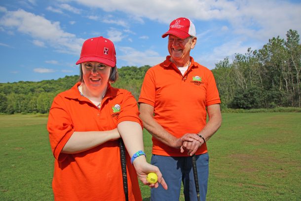 a smiling woman in an orange golf shirt and red ball cap holds a yellow golf ball toward the camera while a smiling man in an orange golf shirt and red ball cap smiles at her.