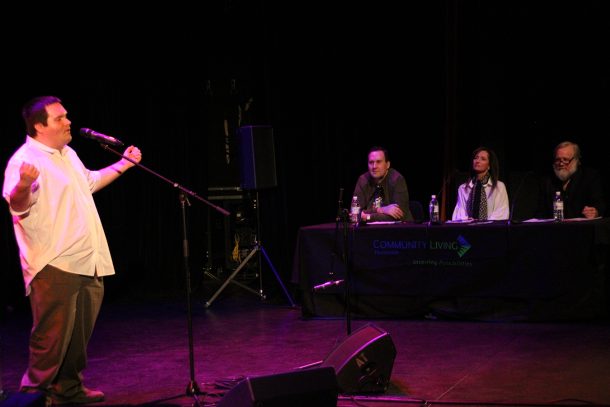 A man in a white shirt sings passionately into a mic on a theatre stage. 3 judges seated at a table nearby watch his performance.