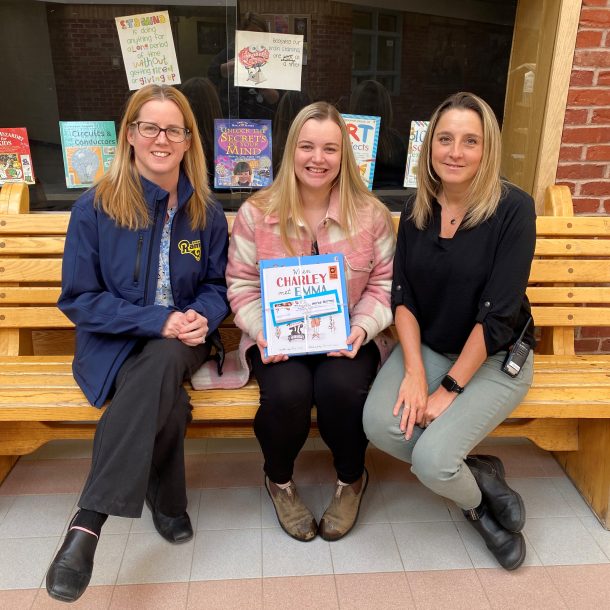 3 smiling woman sit on a bench, indoors. one holds a bundle of books and bookmarks.