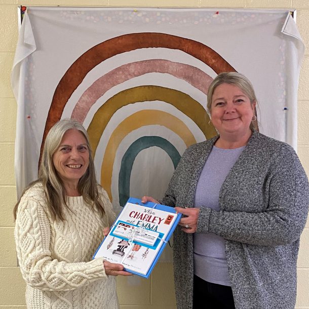 2 smiling women hold a bundle of books and bookmarks. they are standing in front of a rainbow mural.