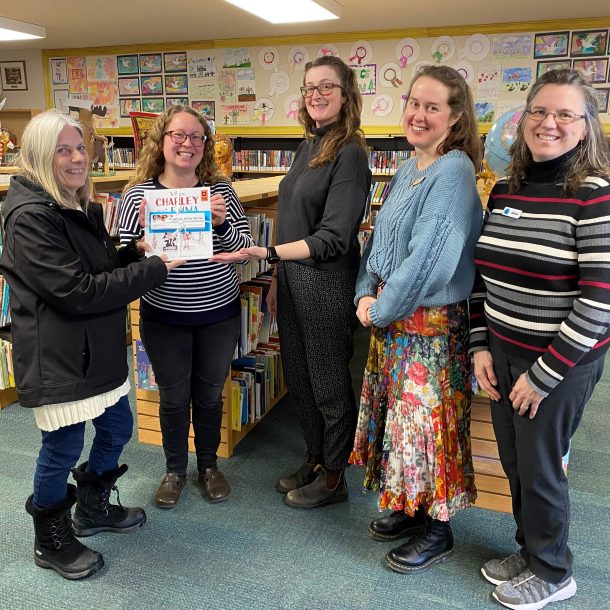 a woman hands a bundle of books and bookmarks to 4 other women. they are standing in the children's section of a library.