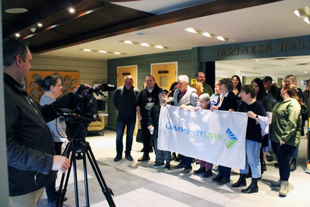 A large group of people gather in a foyer while holding a flag. A TV camera films them.