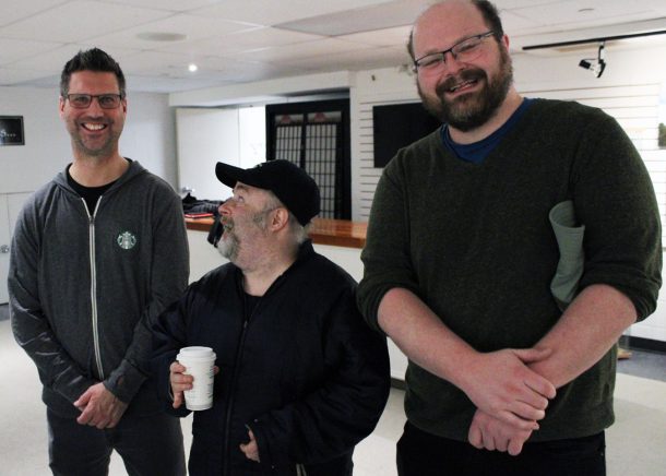 3 smiling men, 1 holding a paper coffee cup.