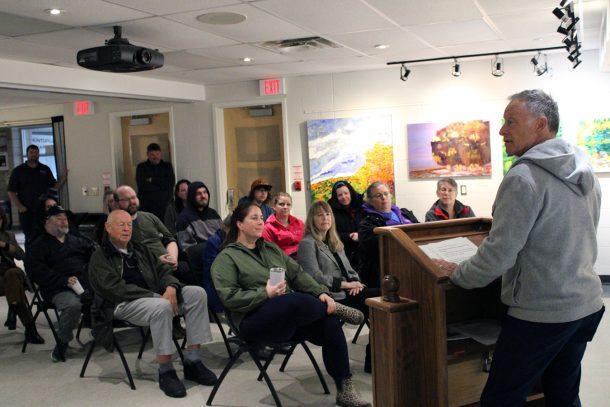 A man at a podium addresses a seated and standing crowd.