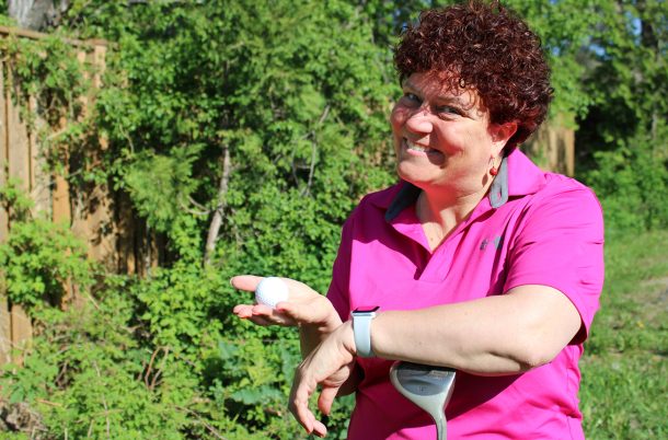 A smiling woman in a pink golf shirt stands outside on a sunny day. She holds a golf ball in one hand, while leaning on a golf club with the other.