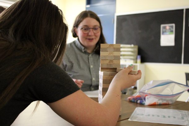 A student pulls a wooden block out of a Jenga tower while an adult, Emily, excitedly looks on.