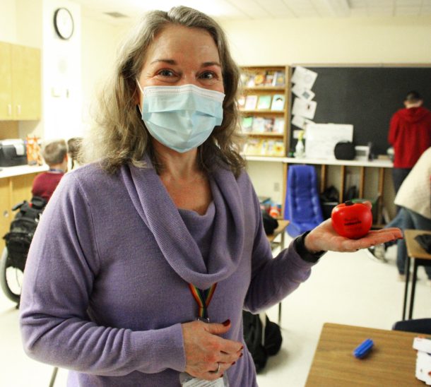 An adult in a face mask, Kim, stands in a classroom holding an apple.