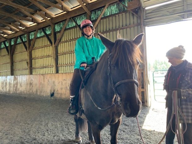 A smiling woman in a green jacket and pink riding helmet on top of a dark brown horse.