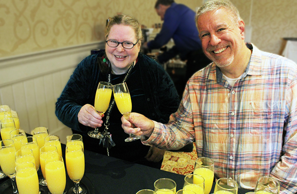 A smiling woman and a smiling man seated at a table while clinking glasses of orange juice together.