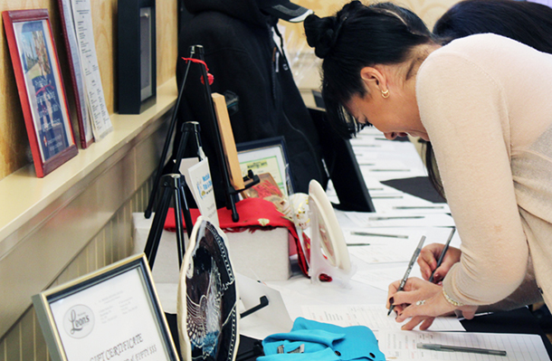 A woman bends over a table to write on a piece of paper. There are silent auction items on the table.