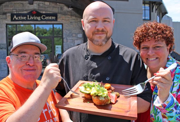 3 smiling people stand outside a building, the Active Living Centre. A man in the centre, a chef, holds a platter of food while the 2 other people stand either side of him holding forks.
