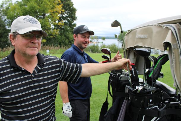 2 men pull golf clubs out of their golf bags at the back of a golf cart.