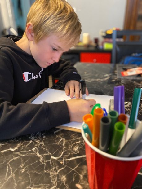 A boy seated at a desk with markers and paper starts to draw a picture.