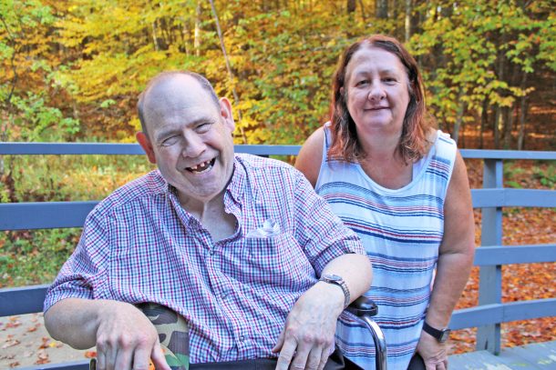 a smiling man and smiling woman pose for a photo on the deck of a house. Nearby trees have colourful fall leaves.