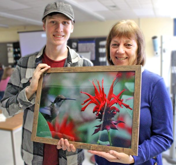 A smiling young man hands a large, framed photograph of a hummingbird on bergamot flowers to a smiling woman.