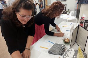 2 women lean over a table to write on silent auction bid sheets.