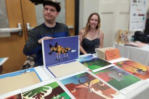 a young man seated at a table covered with art prints. He holds a print of a hyena.