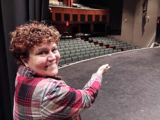 A smiling woman on a performing arts theatre stage gestures with her hand to centre stage.