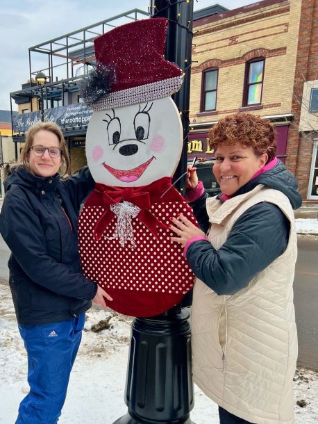 2 smiling women on a downtown main street hold a large wooden cutout shaped and painted like a snowman near a lamp post.