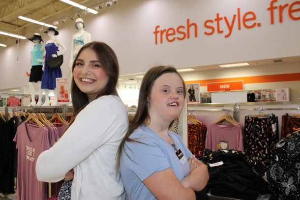 2 smiling young women stand back to back with their arms crossed in a department store's clothing section.