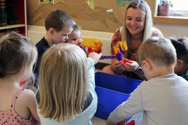 Smiling woman and group of children seated on a classroom floor around a bin of toys.