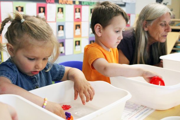 2 children playing with plastic boats that are floating in white tubs filled with water. an adult is nearby.