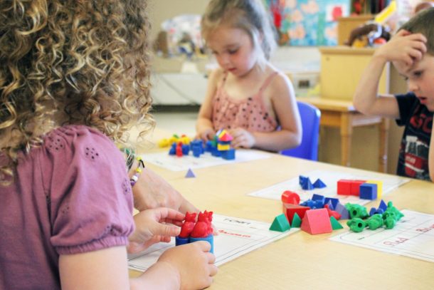 3 children seated at a classroom table stacking small, colourful blocks.