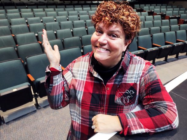 A smiling woman gestures to empty theatre seats behind her.