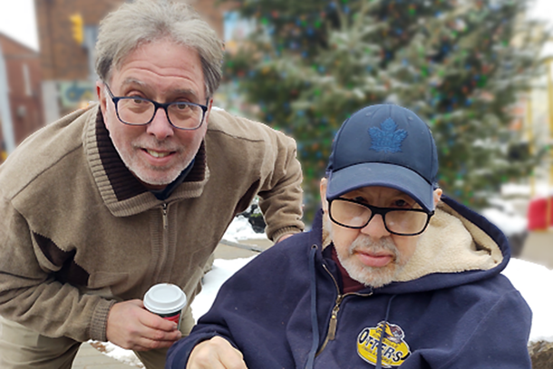 2 smiling men, one standing, one seated in a wheelchair, both holding coffee cups.