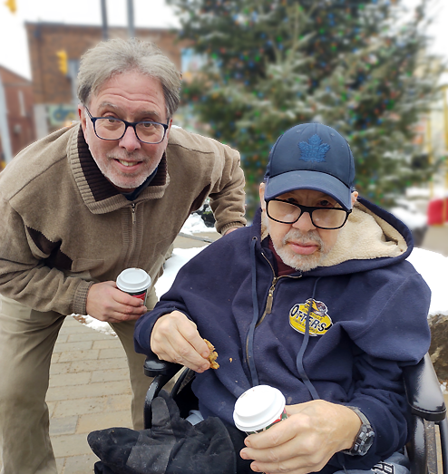2 men, one standing and holding a takeout coffee cup, and one seated in a wheelchair holding a cookie.