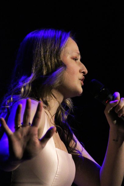 A young woman with blonde hair and a pink dress sings into a microphone while raising her hand.