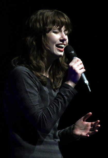 A woman with brown hair sings into a microphone.