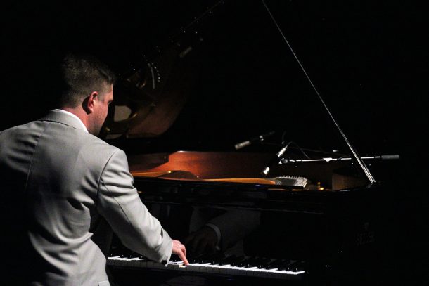 A man in a grey suit jacket plays a grand piano.