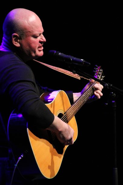 a man in a dark top, John, sings into a microphone while playing the guitar.