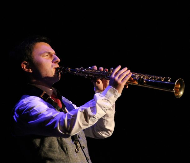 A young man in a white shirt and grey vest, Nils, plays a golden soprano saxophone.