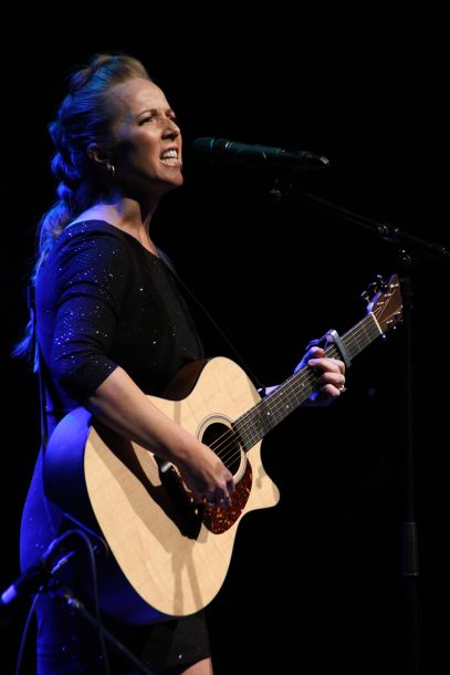 A woman with a long blonde braid, Sonya, sings into a microphone while playing a guitar.