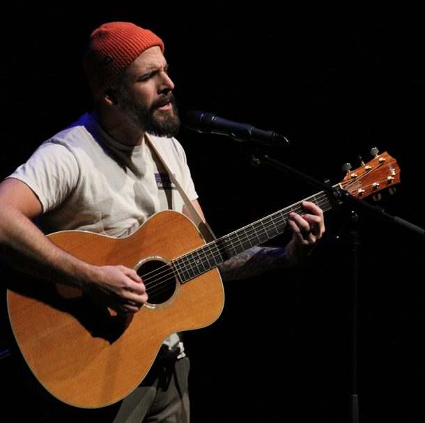 A man in an orange hat and white T-shirt, Todd, signs while playing a guitar.